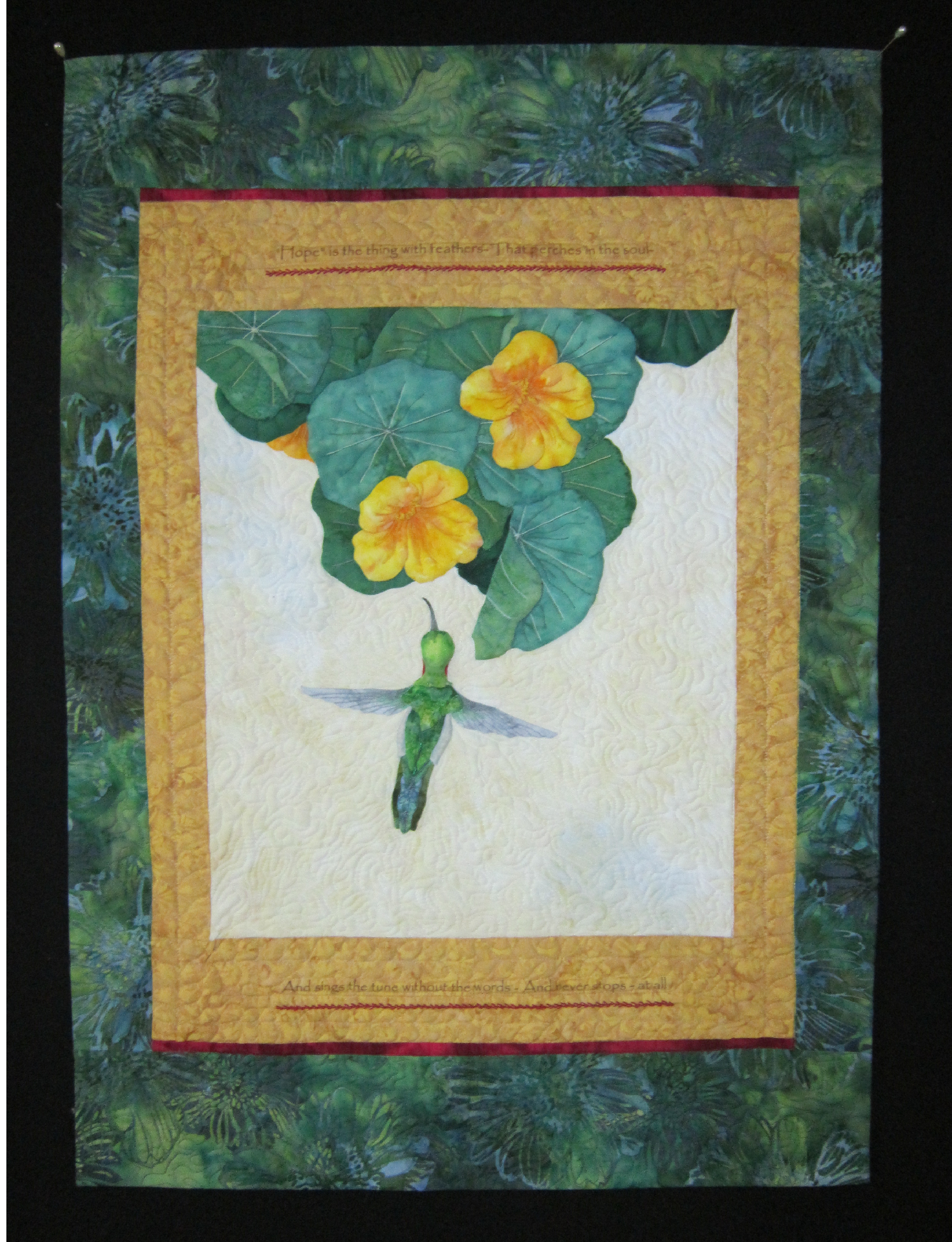 Applique humming bird with flowers and greenery on a cream background with a tan frame on a green background.